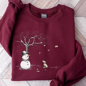 May include: A maroon sweatshirt featuring a winter scene. A snowman wearing a black top hat and scarf stands near a bare tree with snow. A small dog sits nearby, and snowflakes are falling. The sweatshirt is made of a soft material.