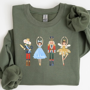 Puede incluir: Una sudadera verde con un diseño festivo que presenta un ratón, una bailarina, un cascanueces y un hada de azúcar. El diseño es de estilo caprichoso con colores brillantes.