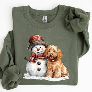 May include: An olive green sweatshirt with a watercolour illustration of a snowman and a golden doodle dog. The snowman wears a hat and scarf. The dog has a red collar. The illustration is set on a white background.