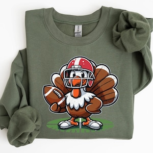 Op de afbeelding: Olijfgroene sweatshirt met een cartoon kalkoen met een footballhelm en een football. De kalkoen is bruin, wit en oranje, met een rood-witte helm. Het ontwerp staat op een groen veld.