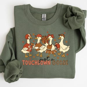 Puede incluir: Sudadera verde oliva con una ilustración de dibujos animados de cuatro gansos vestidos con atuendos de fútbol americano. Los gansos llevan cascos y sostienen un balón y pompones. El texto "just a TOUCHDOWN GOOSE" está impreso debajo.