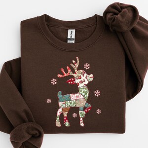 May include: A brown sweatshirt featuring a decorative reindeer design. The reindeer is made of patchwork fabric with red, green, and white patterns. White snowflakes surround the reindeer. The sweatshirt has a crew neck and long sleeves.