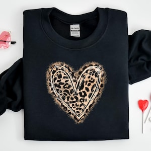 Puede incluir: Sudadera negra con un diseño de corazón de estampado de leopardo. El corazón está contorneado en dorado y tiene un aspecto desgastado.