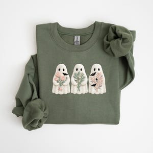 K&ouml;nnte beinhalten: Olivgr&uuml;nes Sweatshirt mit drei wei&szlig;en Geisterfiguren, die Blumen und Flederm&auml;use halten. Ein lang&auml;rmliges Oberteil f&uuml;r Halloween oder den Herbst. Das Design ist ein charmanter, saisonaler Stil.