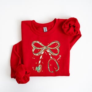 Op de afbeelding: Een rode sweatshirt met een kerstontwerp. De afbeelding bevat een strik en een stethoscoop, beide versierd met zuurstokstrepen en feestelijke kleuren. Het ontwerp is gecentreerd op de voorkant van het shirt met lange mouwen.