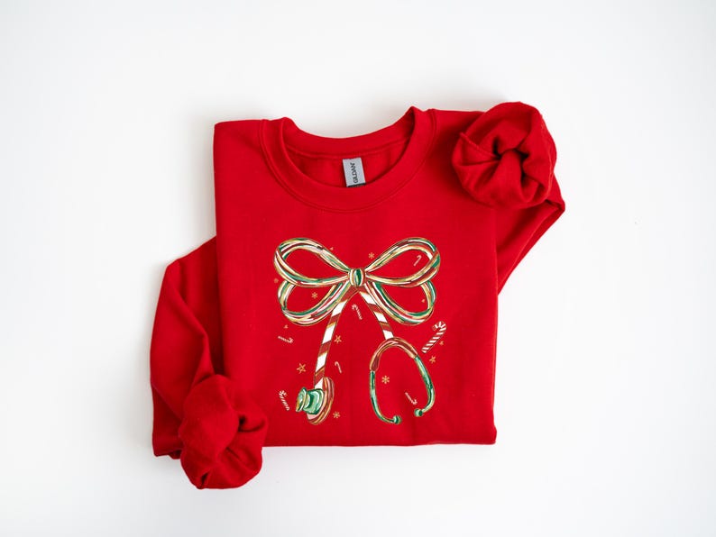 Op de afbeelding: Een rode sweatshirt met een kerstontwerp. De afbeelding bevat een strik en een stethoscoop, beide versierd met zuurstokstrepen en feestelijke kleuren. Het ontwerp is gecentreerd op de voorkant van het shirt met lange mouwen.