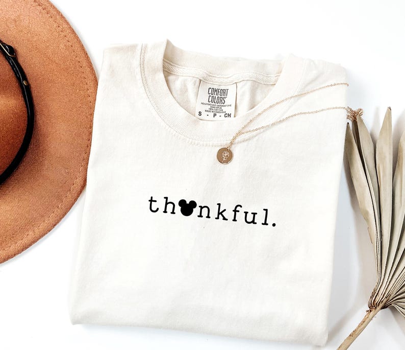 May include: Cream-colored t-shirt with the word "thankful" printed in black, featuring a Mickey Mouse head in place of the "o". A gold necklace and a brown hat are also visible.