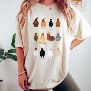 May include: Beige t-shirt featuring a colorful illustration of various chicken breeds. The chickens are depicted in a cartoon style, with detailed plumage and distinct characteristics. The shirt is a relaxed fit, suitable for casual wear.
