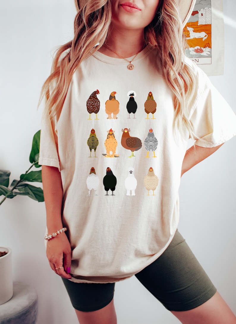 May include: Beige t-shirt featuring a colorful illustration of various chicken breeds. The chickens are depicted in a cartoon style, with detailed plumage and distinct characteristics. The shirt is a relaxed fit, suitable for casual wear.