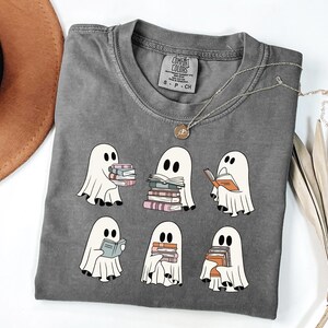 May include: Gray t-shirt featuring a design of six cartoon ghosts reading books. The ghosts are white with black eyes and are holding stacks of books in various colors. The shirt has a crew neck and short sleeves.