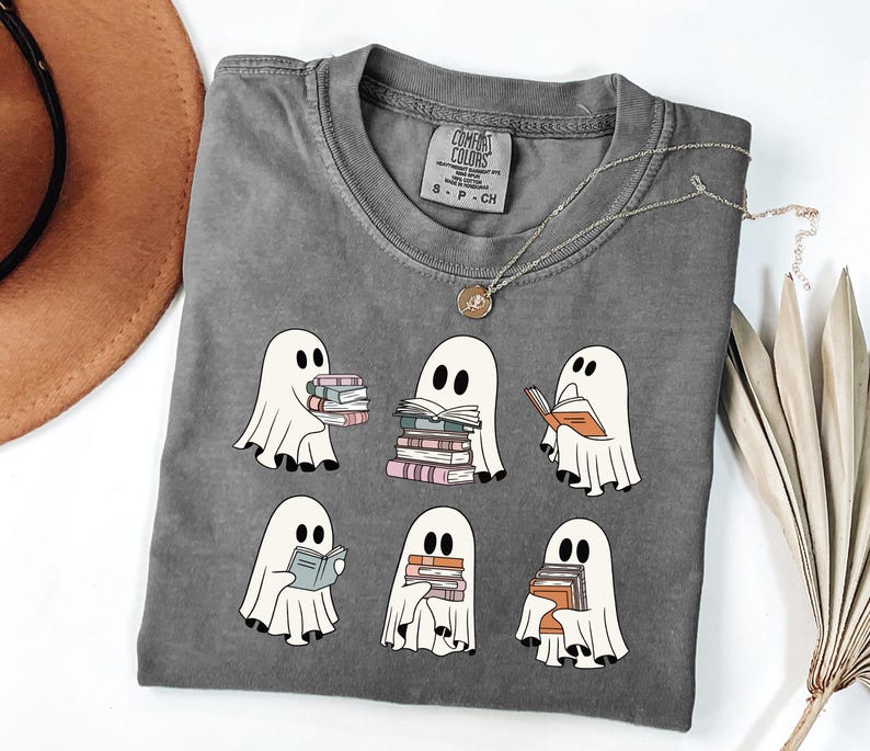 May include: Gray t-shirt featuring a design of six cartoon ghosts reading books. The ghosts are white with black eyes and are holding stacks of books in various colors. The shirt has a crew neck and short sleeves.