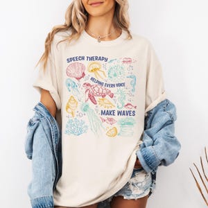 May include: Cream-colored t-shirt with a colorful sea life graphic. The design includes a turtle, jellyfish, seahorse, and seashells, with the text "SPEECH THERAPY HELPING EVERY VOICE MAKE WAVES."
