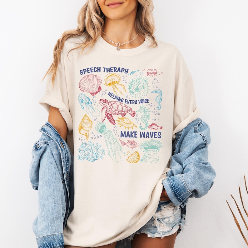 May include: Cream-colored t-shirt with a colorful sea life graphic. The design includes a turtle, jellyfish, seahorse, and seashells, with the text "SPEECH THERAPY HELPING EVERY VOICE MAKE WAVES."