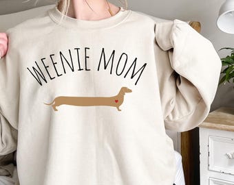 Op de afbeelding: Een crèmekleurig sweatshirt met de tekst "WEENIE MOM" en een bruine teckel met een rood hart op zijn borst.