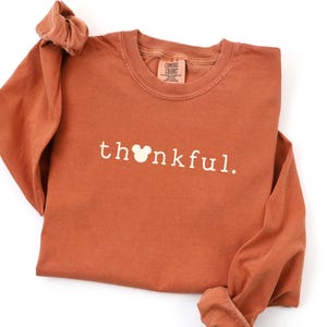 May include: Rust-colored long-sleeve shirt with the word "thankful" printed in white. The "o" in the word is replaced with a Mickey Mouse head. The shirt has a crew neck and the brand "Comfort Colors" is visible on the tag.