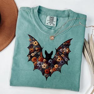 K&ouml;nnte beinhalten: Ein teal-farbenes T-Shirt mit einem floralen Fledermaus-Design. Die Fl&uuml;gel der Fledermaus sind mit bunten Blumen in Orange-, Gelb- und Lilat&ouml;nen gef&uuml;llt. Eine silberne Halskette mit einem kleinen Anh&auml;nger liegt auf dem Shirt.