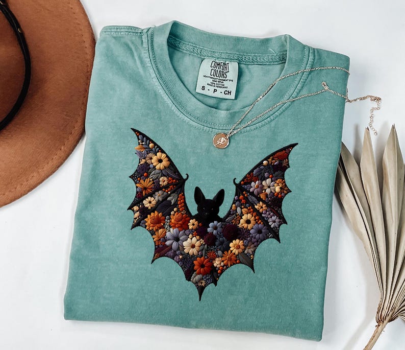 K&ouml;nnte beinhalten: Ein teal-farbenes T-Shirt mit einem floralen Fledermaus-Design. Die Fl&uuml;gel der Fledermaus sind mit bunten Blumen in Orange-, Gelb- und Lilat&ouml;nen gef&uuml;llt. Eine silberne Halskette mit einem kleinen Anh&auml;nger liegt auf dem Shirt.