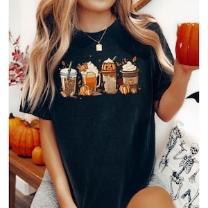May include: Dark grey t-shirt with a graphic of four autumn-themed coffee drinks, pumpkins, leaves, and cinnamon sticks. The shirt has a crew neck and short sleeves.