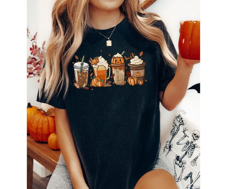 May include: Dark grey t-shirt with a graphic of four autumn-themed coffee drinks, pumpkins, leaves, and cinnamon sticks. The shirt has a crew neck and short sleeves.