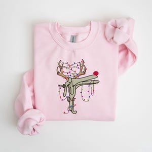May include: A pink sweatshirt with a graphic of a reindeer with Christmas lights wrapped around its antlers. The reindeer is wearing a red nose.