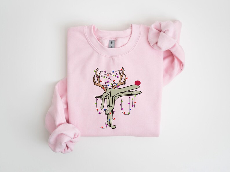 May include: A pink sweatshirt with a graphic of a reindeer with Christmas lights wrapped around its antlers. The reindeer is wearing a red nose.