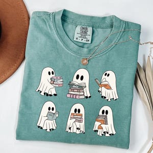 May include: A teal Comfort Colors t-shirt featuring a graphic of six cartoon ghosts reading books. The ghosts are white with black eyes and are holding stacks of books in various colors. A gold necklace with a pendant is draped on the shirt.