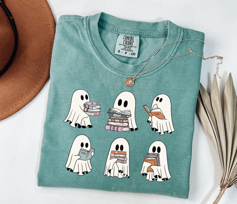 May include: A teal Comfort Colors t-shirt featuring a graphic of six cartoon ghosts reading books. The ghosts are white with black eyes and are holding stacks of books in various colors. A gold necklace with a pendant is draped on the shirt.