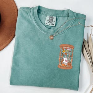 May include: A seafoam green t-shirt with a round neckline and short sleeves. The shirt features a graphic of an hourglass with a ghost, pumpkins, and autumn leaves. A gold necklace with a pendant rests on the shirt.