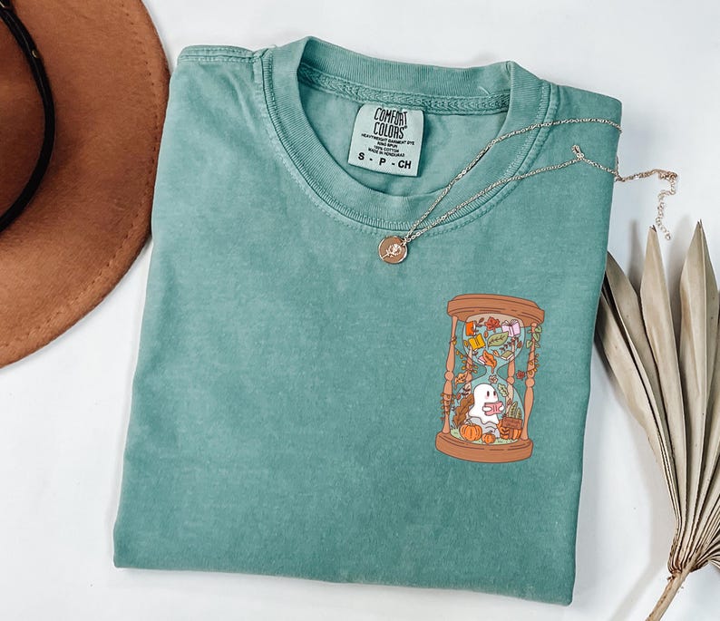 May include: A seafoam green t-shirt with a round neckline and short sleeves. The shirt features a graphic of an hourglass with a ghost, pumpkins, and autumn leaves. A gold necklace with a pendant rests on the shirt.