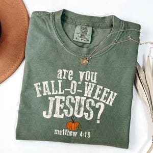 Puede incluir: Camiseta verde oliva con el texto "are you FALL-O-WEEN JESUS?" y un gráfico de calabaza. La camiseta también presenta el versículo bíblico Mateo 4:19. Se ve un collar dorado y un sombrero marrón.