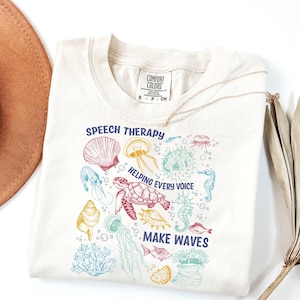 Puede incluir: Camiseta color crema con un gráfico colorido de vida marina. El diseño incluye una tortuga, medusas, un caballito de mar y varias conchas. El texto dice "Speech Therapy Helping Every Voice Make Waves."