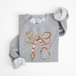 Op de afbeelding: Een grijze sweatshirt met een feestelijk ontwerp van een stethoscoop gemaakt van zuurstokken. De stethoscoop heeft de vorm van een strik, met rode, groene en witte strepen. Zuurstokken zijn verspreid rond het ontwerp.