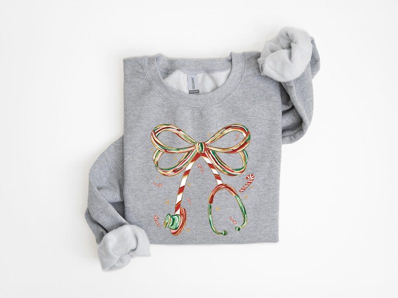 Op de afbeelding: Een grijze sweatshirt met een feestelijk ontwerp van een stethoscoop gemaakt van zuurstokken. De stethoscoop heeft de vorm van een strik, met rode, groene en witte strepen. Zuurstokken zijn verspreid rond het ontwerp.