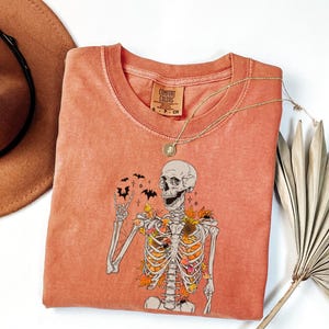 May include: Coral-colored Comfort Colors t-shirt featuring a skeleton graphic with autumn leaves and bats. The skeleton is giving a peace sign. A gold necklace and a brown hat are also in the image.