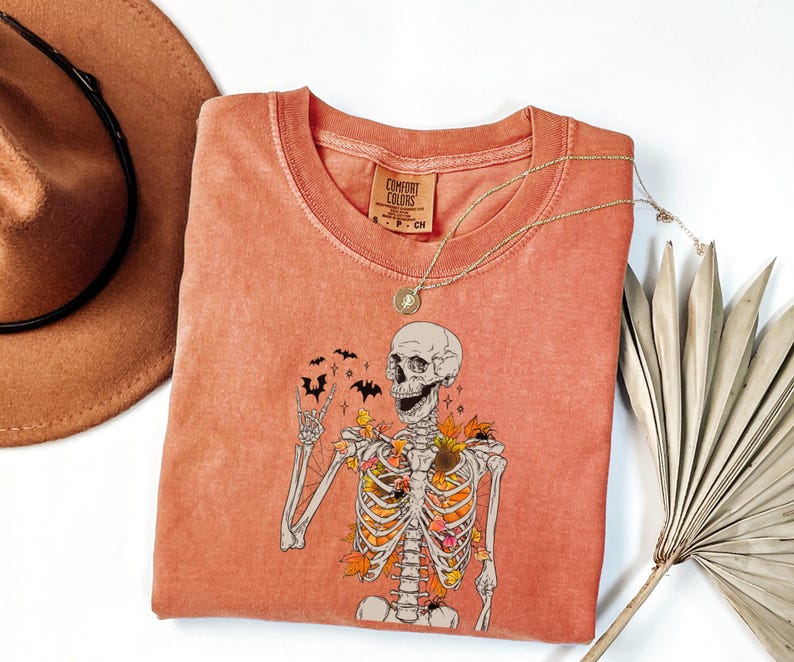 May include: Coral-colored Comfort Colors t-shirt featuring a skeleton graphic with autumn leaves and bats. The skeleton is giving a peace sign. A gold necklace and a brown hat are also in the image.