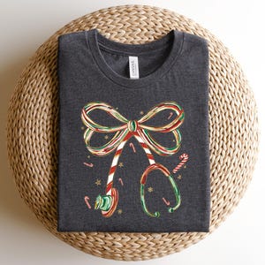 Op de afbeelding: Donkergrijs t-shirt met een kerstthema. De afbeelding bevat een strik en een stethoscoop, beide versierd met zuurstokstrepen en accenten. Het ontwerp bevat ook zuurstokken en kleine sterren.