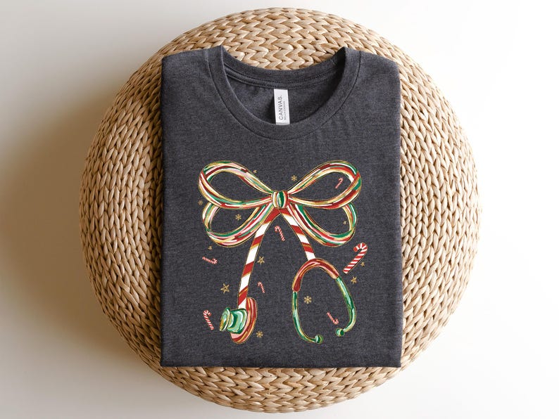 Op de afbeelding: Donkergrijs t-shirt met een kerstthema. De afbeelding bevat een strik en een stethoscoop, beide versierd met zuurstokstrepen en accenten. Het ontwerp bevat ook zuurstokken en kleine sterren.