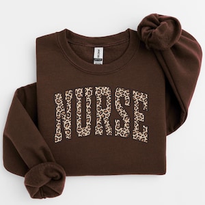Puede incluir: Una sudadera marrón de cuello redondo con la palabra "NURSE" en letras grandes con estampado de leopardo en el pecho. La sudadera tiene mangas largas y cuello y puños acanalados. La etiqueta de la marca Gildan es visible.