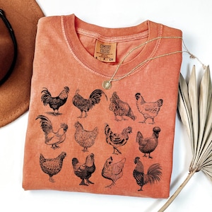 May include: Coral-colored Comfort Colors t-shirt featuring a collection of black rooster and hen illustrations. A gold necklace with a pendant rests on the shirt. The shirt is folded, with a hat and decorative plant in the background.