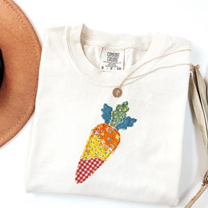Può includere: T-shirt color crema con applicazione a carota. Il design della carota presenta tessuti arancioni, gialli e rossi con motivi floreali e a quadri. Una collana d'oro con un piccolo ciondolo poggia sulla maglietta. La maglietta ha un'etichetta Comfort Colors.