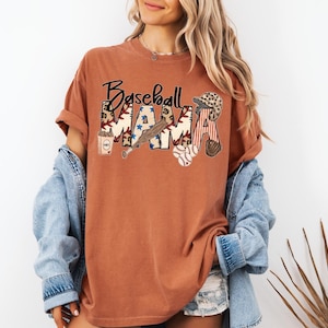 May include: A rust-colored t-shirt with the words "Baseball Mama" in a decorative font. The word "MAMA" is filled with baseball-themed graphics, including stars, a baseball bat, and a baseball glove. The shirt has short sleeves and a relaxed fit.