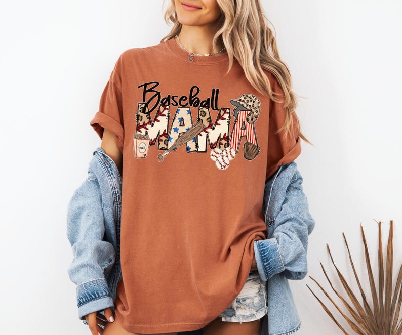 May include: A rust-colored t-shirt with the words "Baseball Mama" in a decorative font. The word "MAMA" is filled with baseball-themed graphics, including stars, a baseball bat, and a baseball glove. The shirt has short sleeves and a relaxed fit.