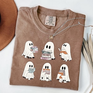 May include: A brown Comfort Colors t-shirt featuring a graphic of six white ghosts reading books. The ghosts have black eyes and are holding stacks of books in various colors. A silver necklace with a pendant is also visible.