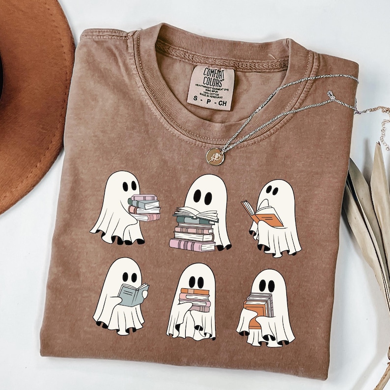May include: A brown Comfort Colors t-shirt featuring a graphic of six white ghosts reading books. The ghosts have black eyes and are holding stacks of books in various colors. A silver necklace with a pendant is also visible.