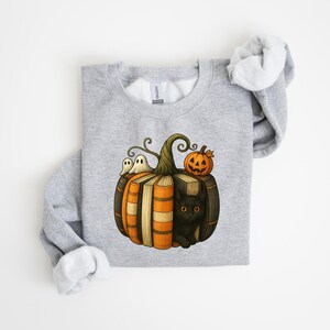 May include: A light gray sweatshirt featuring a Halloween-themed graphic. The design includes a pumpkin made of books, a black cat, two ghosts, and a jack-o'-lantern. The sweatshirt has a crew neck and long sleeves.