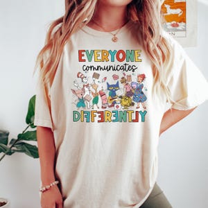 May include: A cream-colored t-shirt with the text "EVERYONE communicates DIFFERENTLY" in rainbow letters. The shirt features a colorful illustration of characters from popular children's books, including Cat in the Hat, Clifford the Big Red Dog, and The Very Hungry Caterpillar.