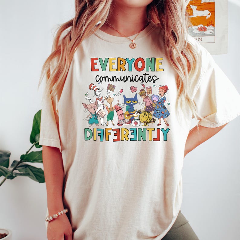 May include: A cream-colored t-shirt with the text "EVERYONE communicates DIFFERENTLY" in rainbow letters. The shirt features a colorful illustration of characters from popular children's books, including Cat in the Hat, Clifford the Big Red Dog, and The Very Hungry Caterpillar.