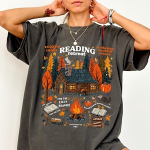 May include: Dark gray t-shirt with a colorful graphic featuring a cabin in a forest setting. The design includes text that reads "Reading Retreat," "Cozy Cabin," and "For the Cozy Readers." The shirt has a relaxed fit.