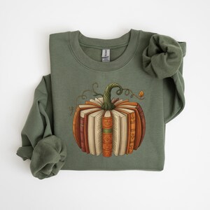 May include: Olive green sweatshirt featuring a pumpkin design made of books. The pumpkin has a green stem and is composed of various books in shades of brown, orange, and cream. The design is centered on the front of the long-sleeved top.