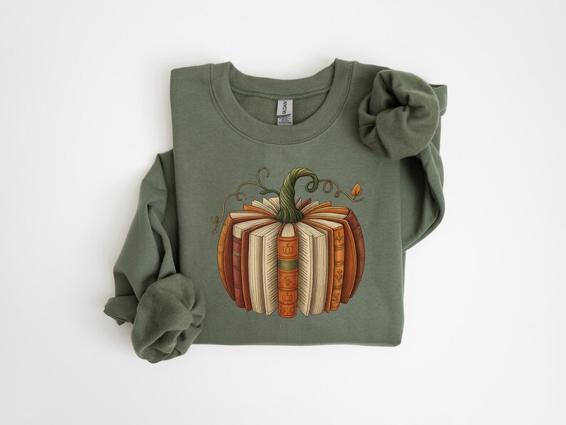 May include: Olive green sweatshirt featuring a pumpkin design made of books. The pumpkin has a green stem and is composed of various books in shades of brown, orange, and cream. The design is centered on the front of the long-sleeved top.